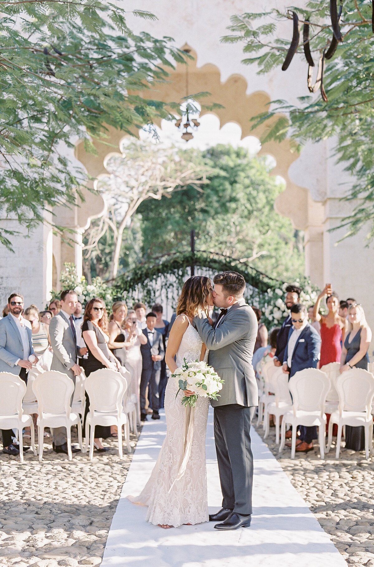 A romantic outdoor wedding ceremony at a top tropical destination wedding venue in the Caribbean, Dominican Republic captured by photographer Laura Gordon. A couple shares a kiss at the end of the aisle, surrounded by friends and family, with the bride in a lace gown holding a bouquet and the groom in a gray suit. They stand under an elegantly decorated arch with lush greenery.