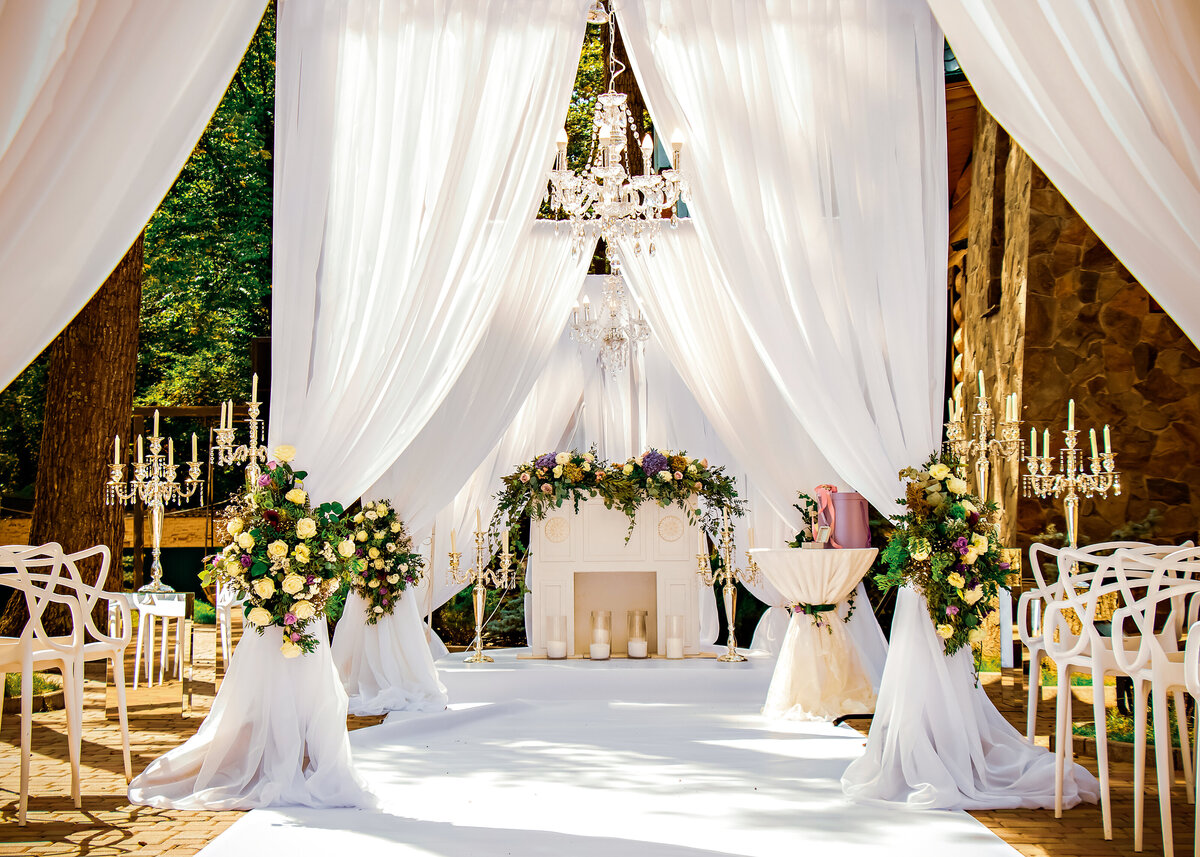 Wedding ceremony decor, with white fireplace and chandeliers decorated with flowers, white drapes and white chairs outdoors.