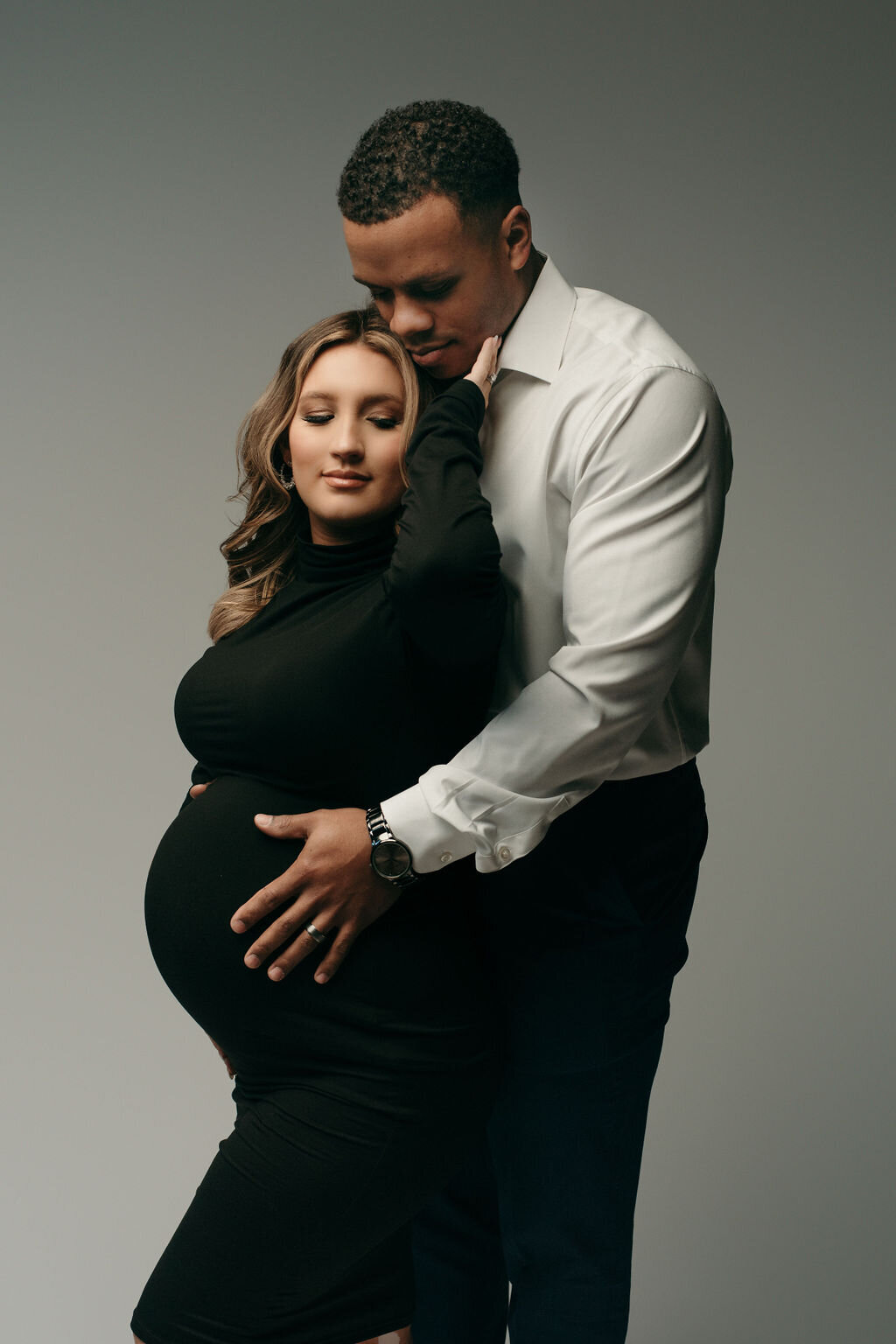 Man and woman posing for maternity portrait with man standing behind woman holding her baby bump