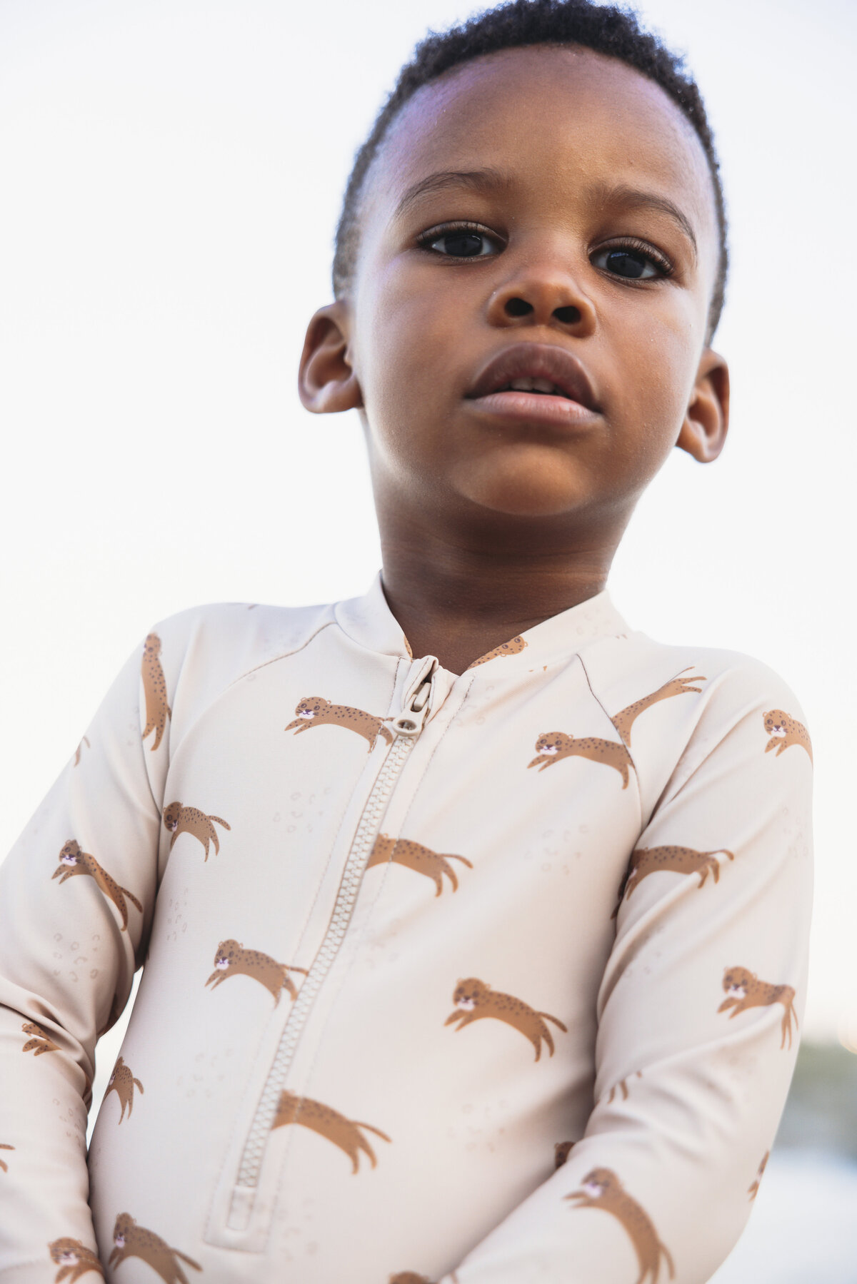 Close-up portrait of a young boy wearing a beige swim shirt with playful leopard prints, looking into the camera with a calm expression.