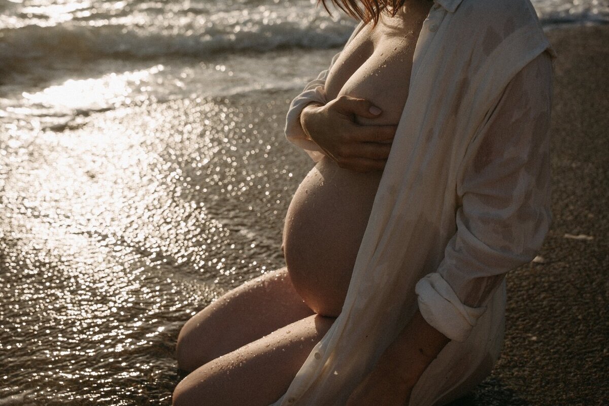 Femme enceinte agenouillée sur le sable humide, chemise ouverte laissant apparaitre son ventre. Séance intime et naturelle par Kamescop, photographe maternité.