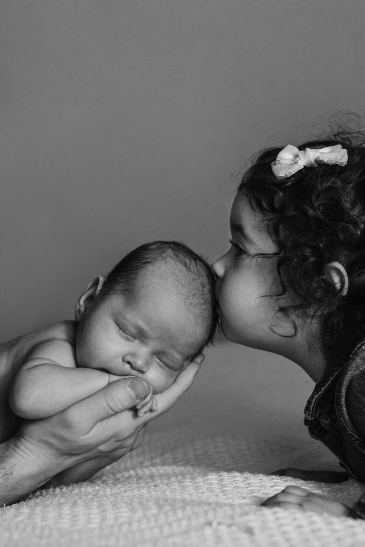 Toddler sister kisses new baby brother on the head while dad hols him black and white Newborn Portrait Photography