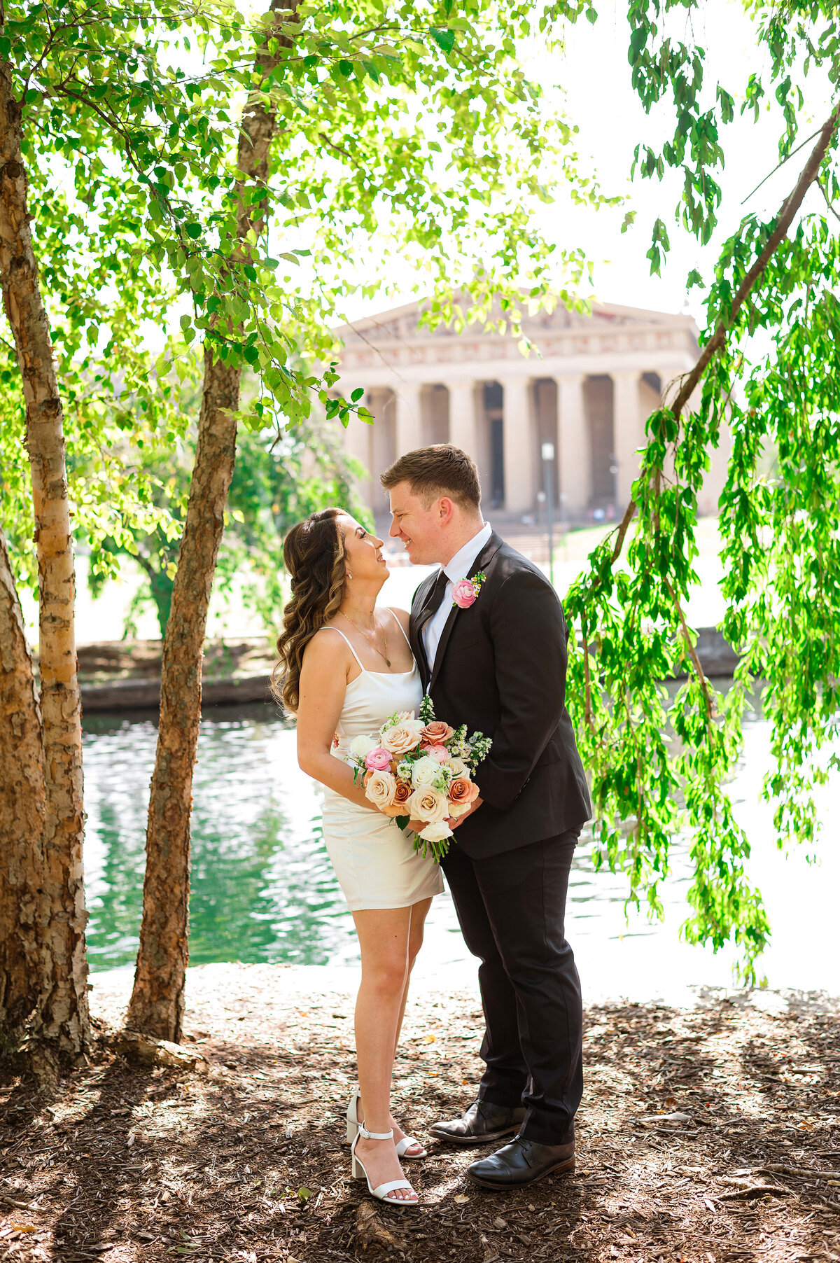 Bride and groom smiling at each other under willow trees with Nashville Parthenon behind them