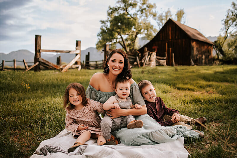 om holds her three children on her lap while posing for a photo in front of a brown barn