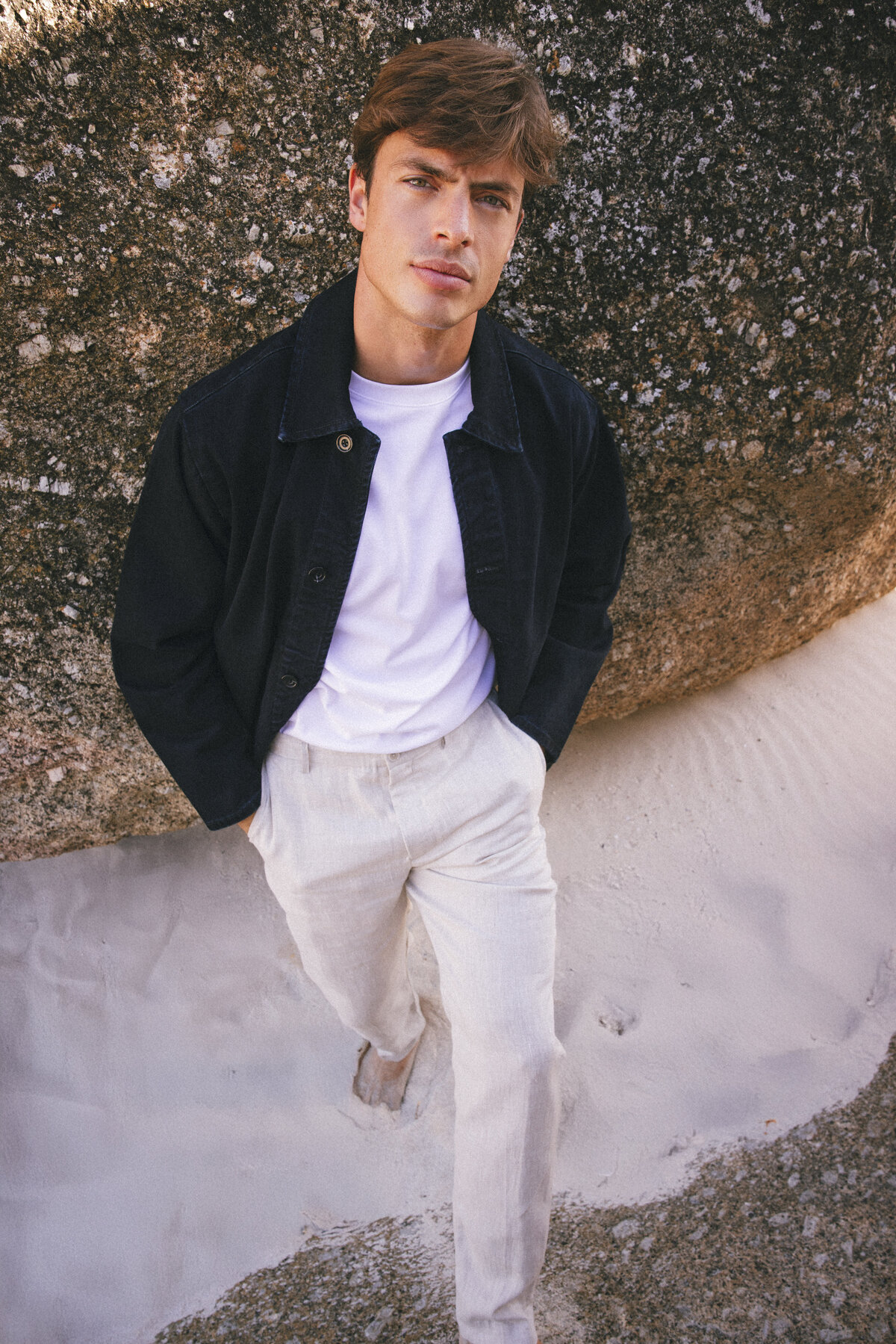 Male model standing barefoot on white sand against a textured rock, wearing a white T-shirt, light linen trousers, and a dark overshirt. Shot in Cape Town, the image conveys a relaxed and refined summer aesthetic.