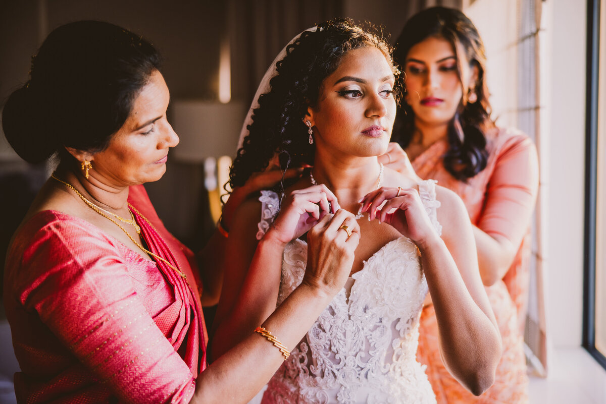 An indian malayali christian bride getting ready for her wedding with the help of her mother and sister in front of a window at a hotel