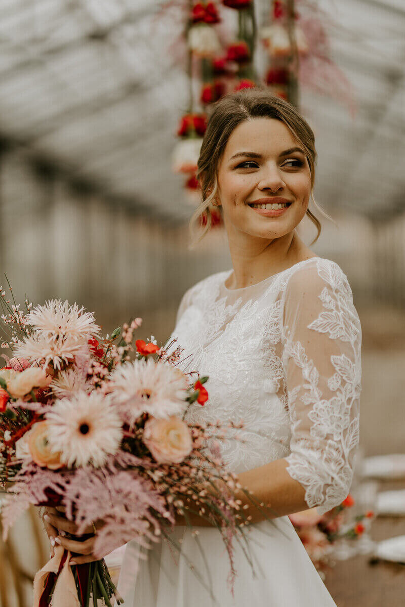 Mariée de profil tenant son bouquet rouge et rose regard vers la droite posant sous une verrière devant une table décorée pour un mariage. Shooting photo pour le workshop photographie de mariage.