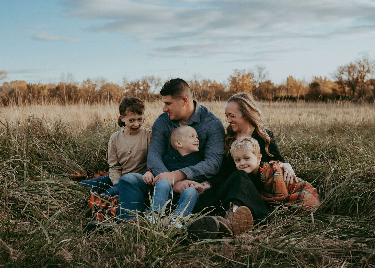 A mom and dad sit on a blanket during fall with their three wild boys, laughing and giggling while wearing a stylish neutral color palette that coordinates perfectly with the bottom tones of the tall grasses they're sitting in.