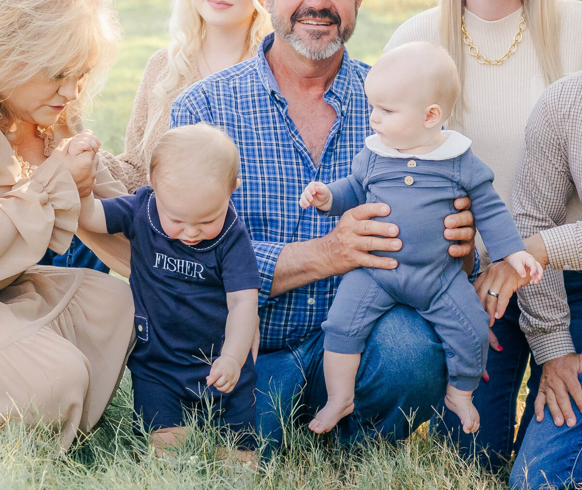 toddler picking grass while baby cousin watches him while grandparents are holding them