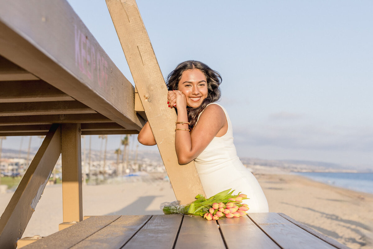 A woman in a white dress leans against a lifeguard tower on a sunny beach. She gazes at the camera, with a bouquet of tulips resting on a wooden table in the foreground. The ocean and clear sky form the backdrop.