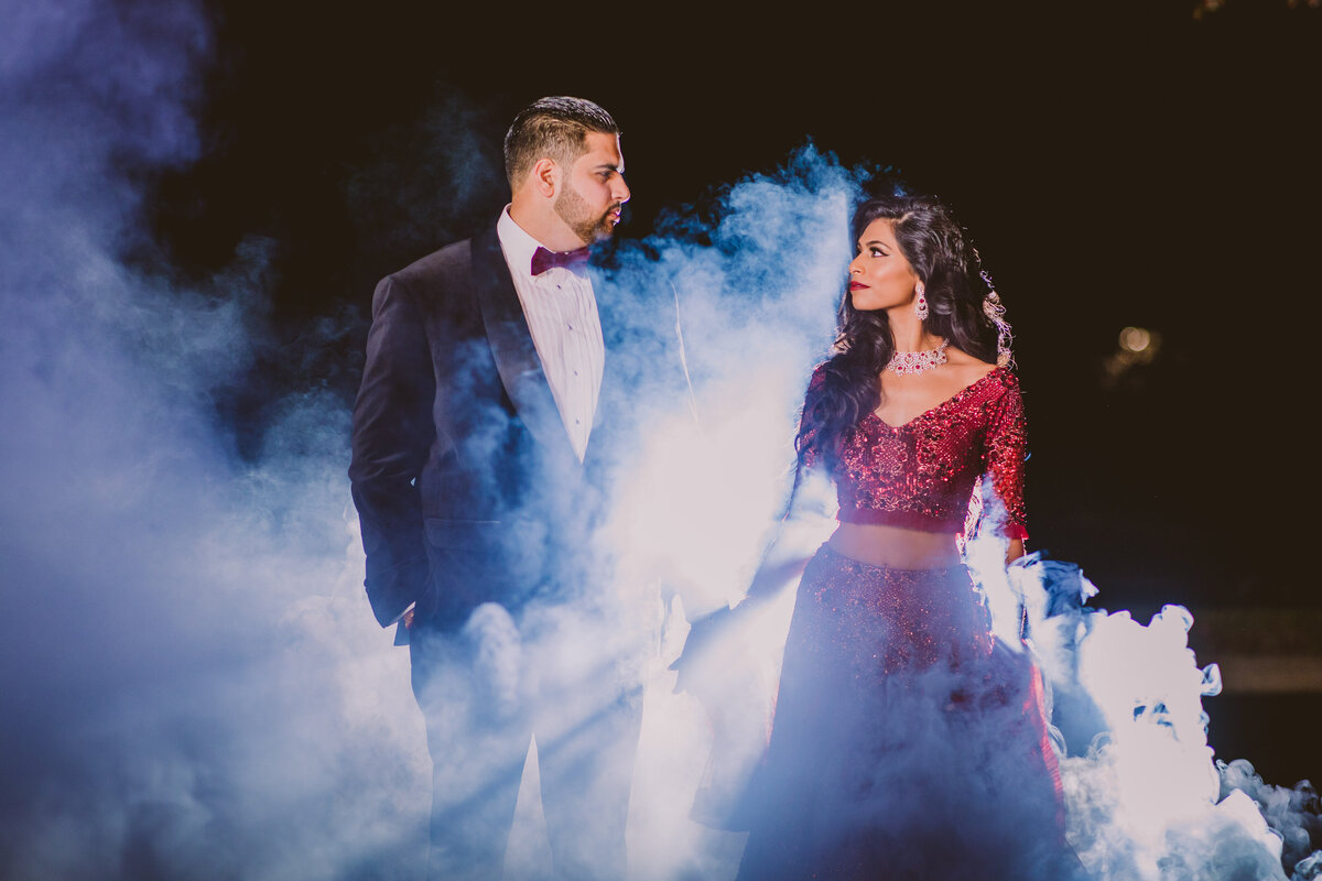 indian bride and groom looking at each other outside at night with a white smoke bomb smoke around them