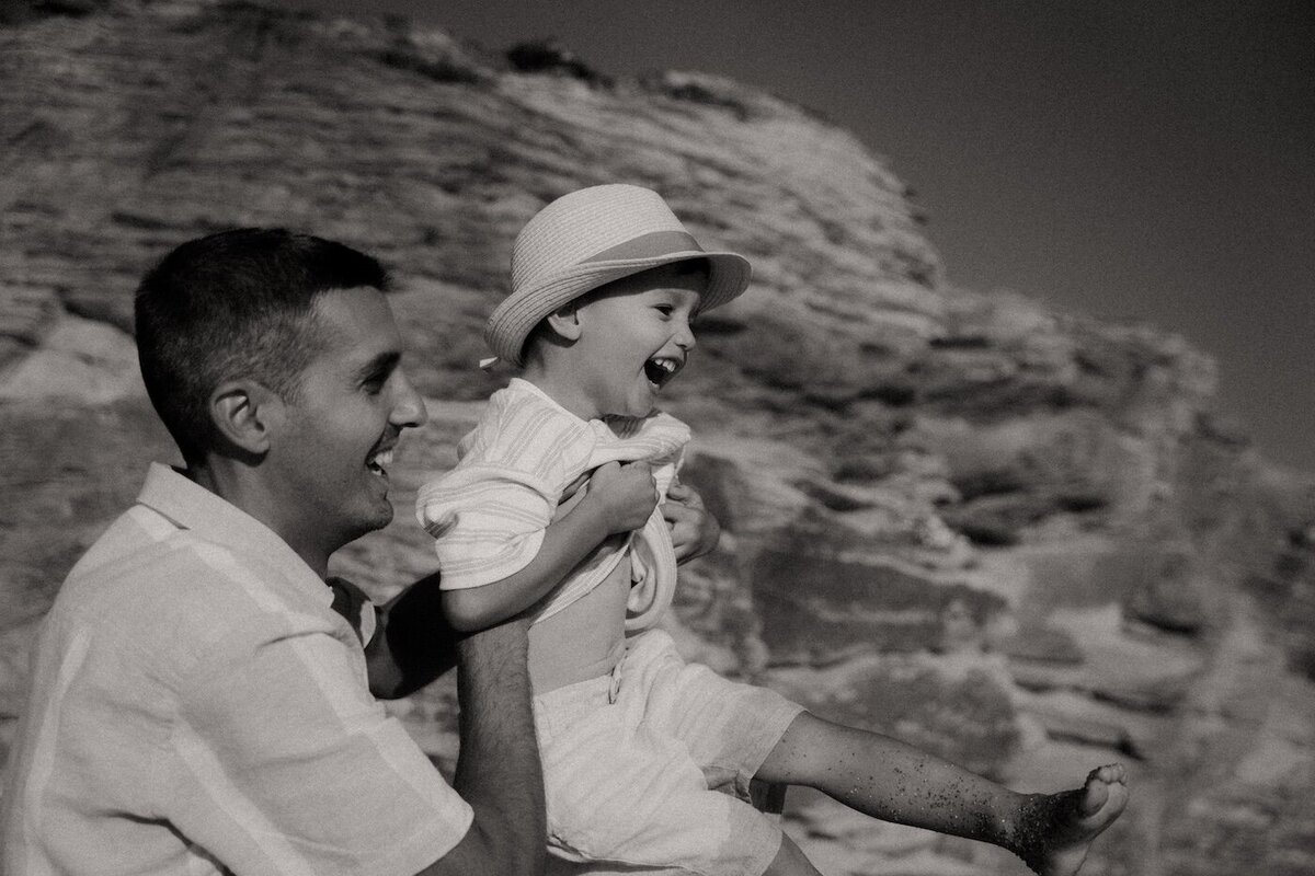 Le père porte son fils dans ses bras. L'enfant rit, les jambes pleines de sable. Photo réalisée par Kamescop, photographe famille à Pornic.