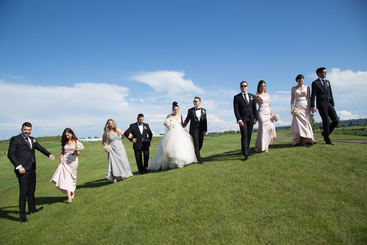 The bridal party poses together in a lush, open field, surrounded by greenery. The bride and groom are at the center, flanked by their attendants dressed in formal attire. The natural setting adds a relaxed and picturesque feel to the group's wedding portrait, highlighting the joy and unity of the special day.