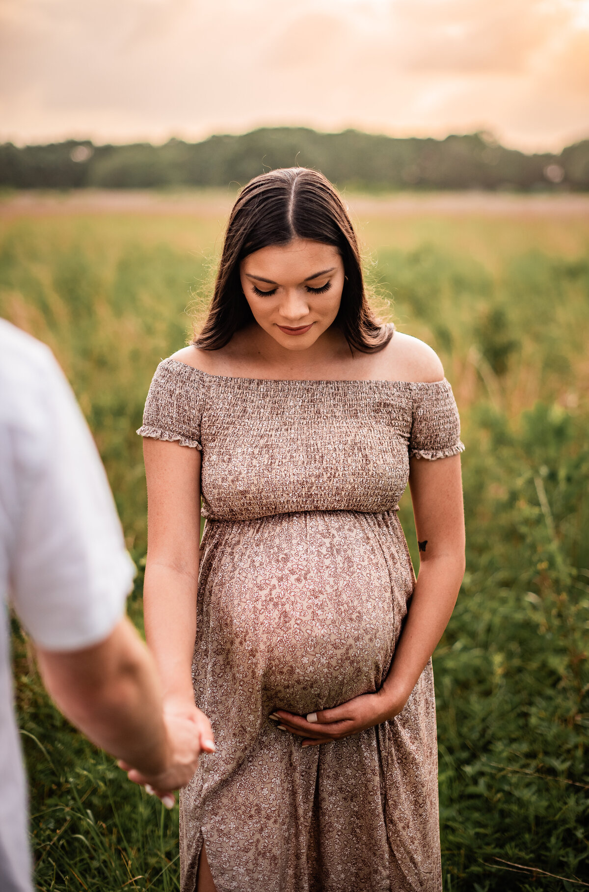 A woman holds her baby bump and looks down at er belly while her boyfriend holds her hand in the foreground.