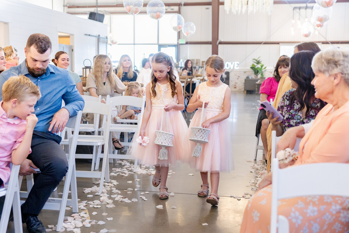  Two young flower girls in pastel pink dresses walk down the aisle carrying macrame flower baskets, scattering petals with a sweet innocence. Guests look on as they set the stage for the bride’s grand entrance. 