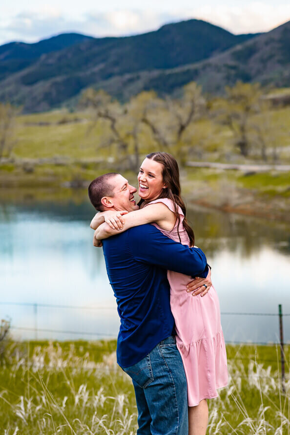 A man picks up his wife as they both laugh, with a lake in the background.