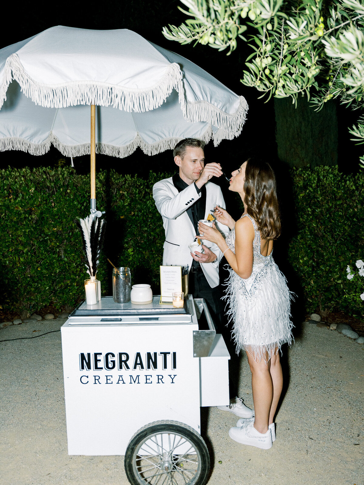 A bride and groom feed each other ice cream during their wedding reception at Alegrehtto Vineyard she is wearing a second wedding dress covered with sequence and feathers