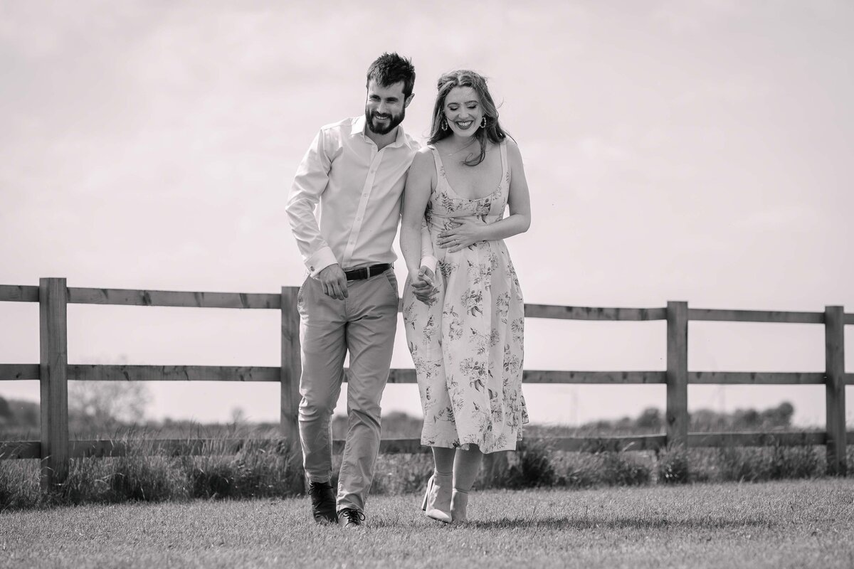 A black and white photo of an engaged couple walking through a meadow hand in hand