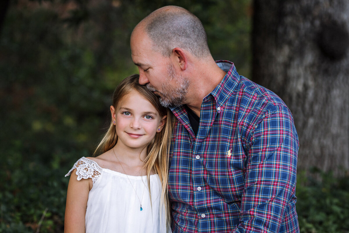 8-year-old girl sitting next to her dad at Bay Area park as he leans over to kiss the top of her head