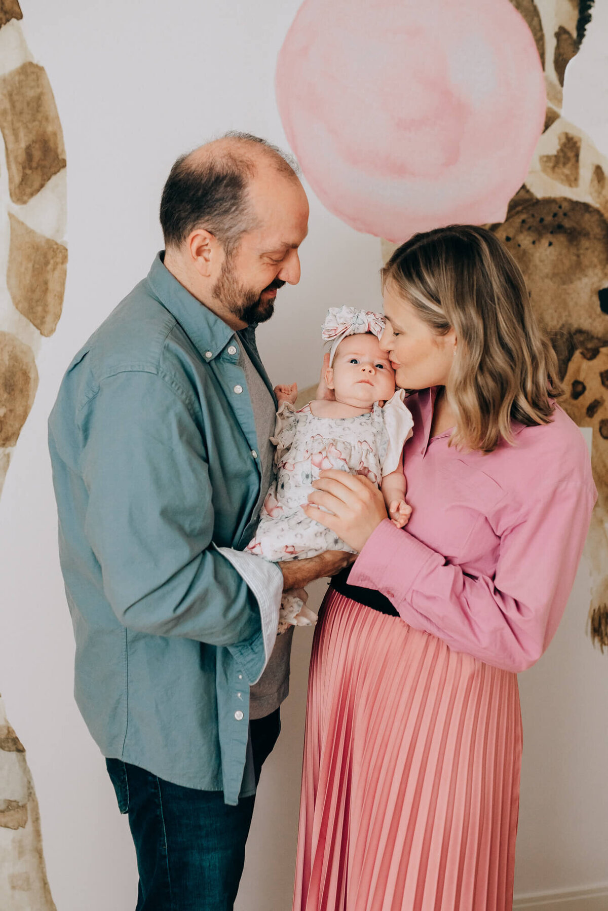 A mother and father hold their newborn daughter together as the mother gives her a kiss in her safari themed nursery