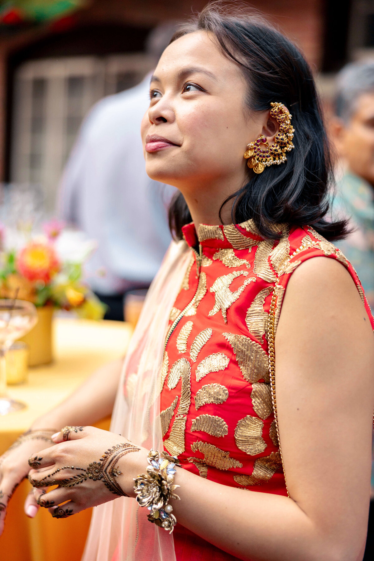 A woman wearing a red and gold embroidered dress and ornate earrings stands with her hands adorned with floral jewelry and intricate henna. She appears to be at a celebratory event with soft-focus floral decorations in the background.