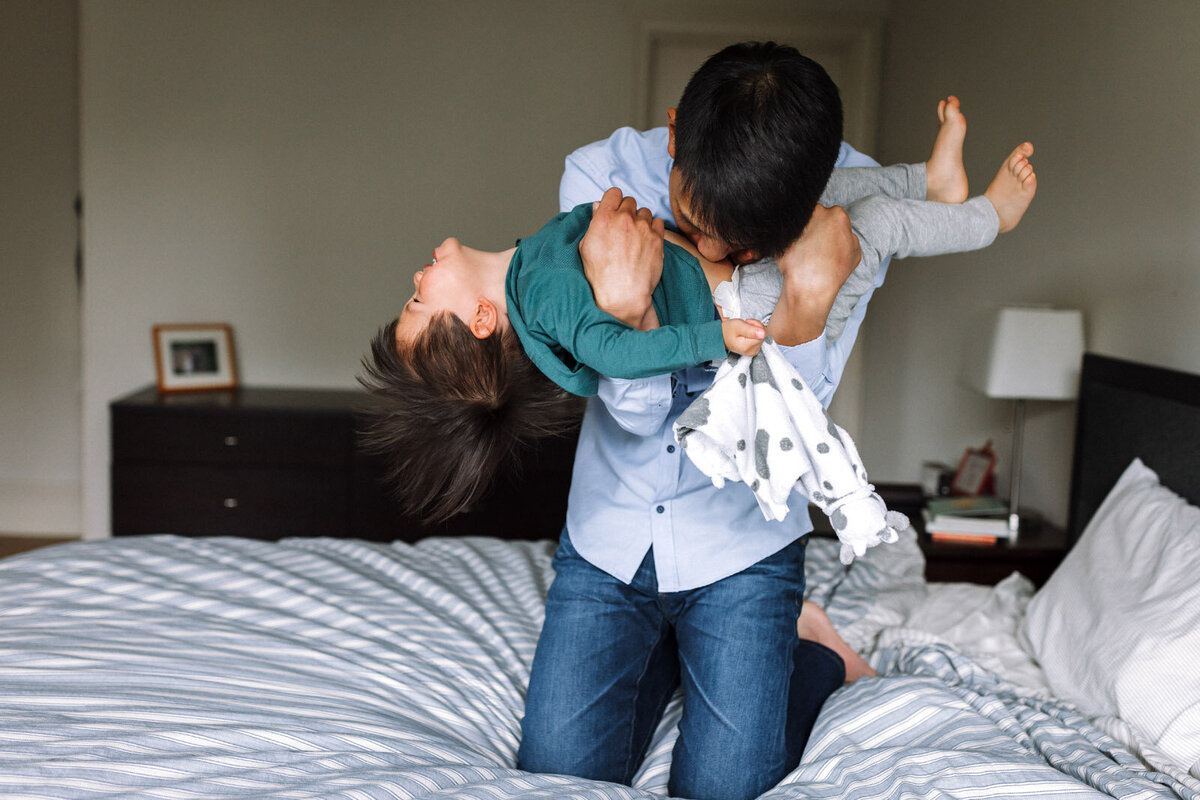 dad holding young son on bed and giving him a raspberry on his stomach as the child laughs