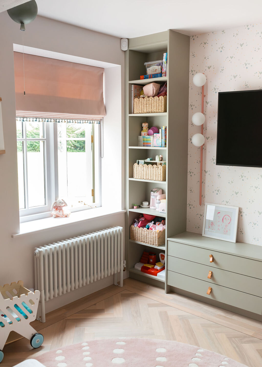 A cozy children's playroom with pastel-colored decor. The walls are decorated with patterned wallpaper and a flat-screen TV is mounted on the wall. To the left is a window and shelves filled with toys and baskets.