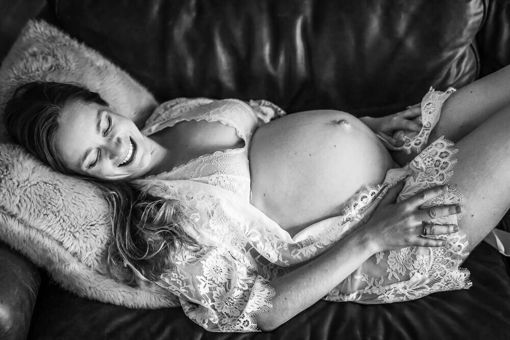 A black and white image of a first time mother in a lacy robe, lounging on her couch during a boudoir style Maternity in home photo session.