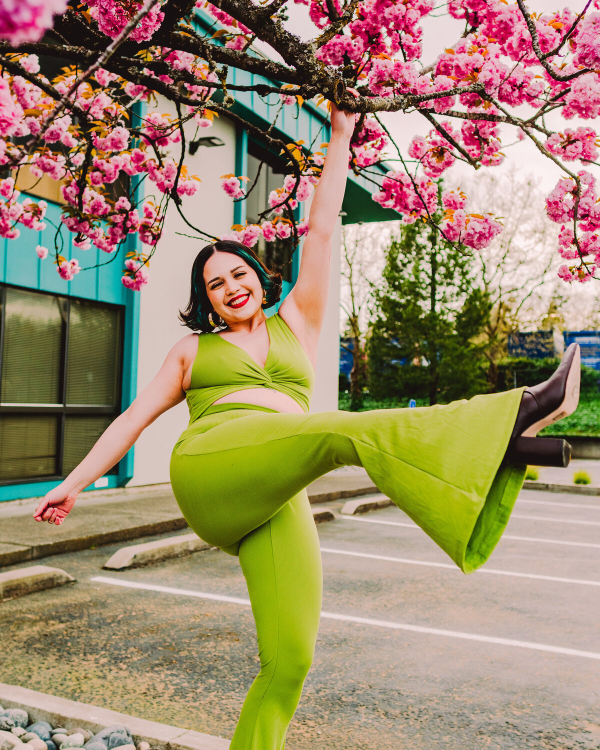 Creative woman in a vibrant green jumpsuit hanging from a blossoming cherry tree, smiling brightly – joyful outdoor brand photo by Mewphotovideo