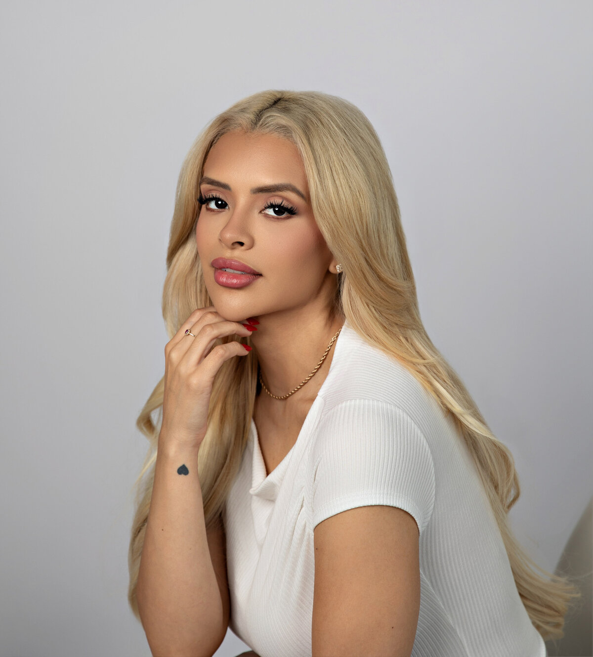 Studio portrait of a blonde woman in a white top, hand resting gently on chin, with soft makeup and a poised expression—Eva Magaña Photography.