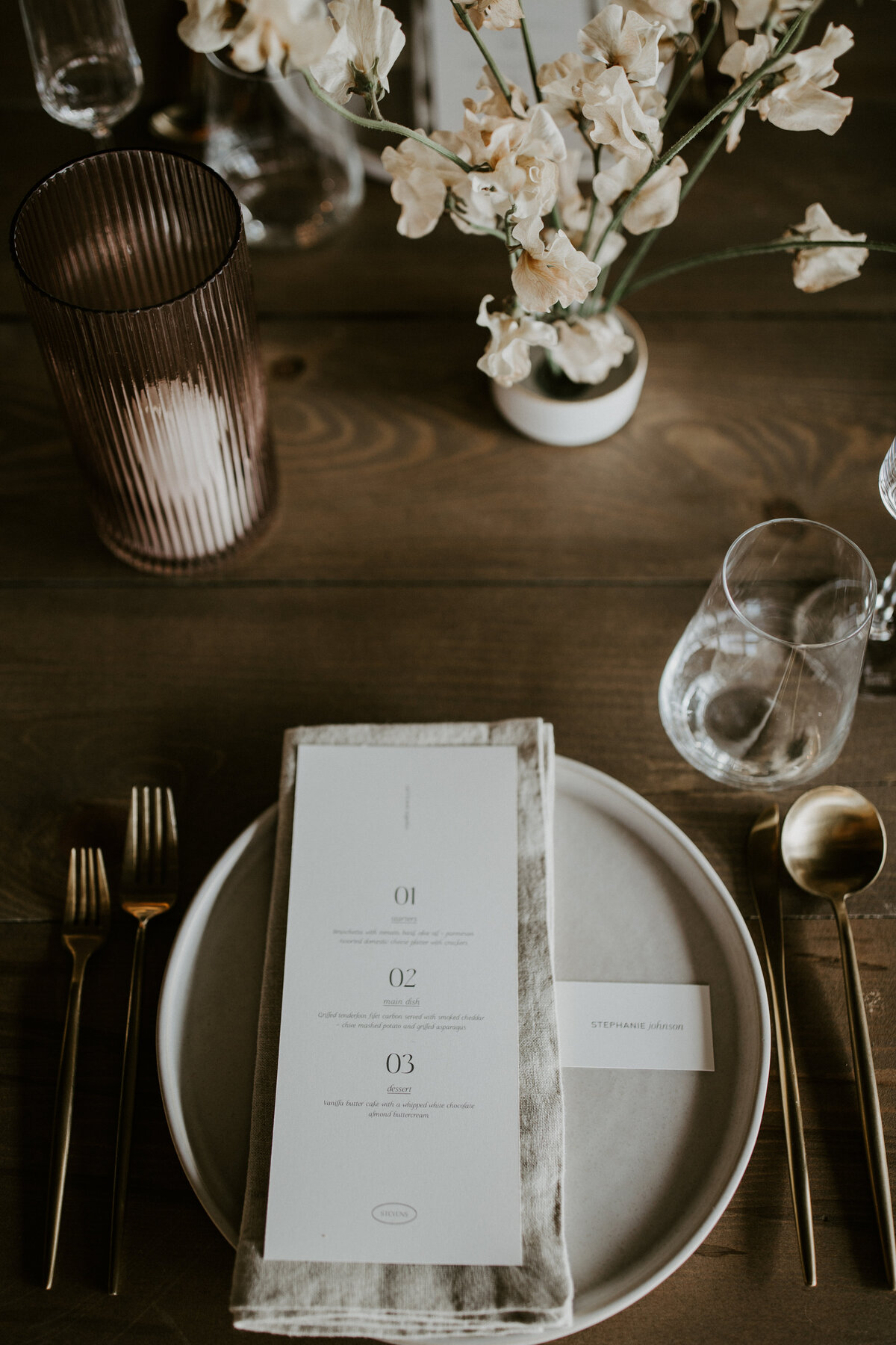 White dinner menu with black font atop a white linen napkin and white plate set on wooden table with gold silverware.