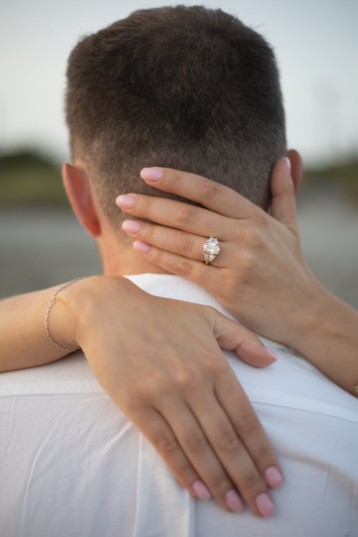 Collins Beach Newport RI | Kelsey Sheehan Photography Timeless Rhode Island Weddings | A woman's hands gently embrace a man's neck from behind, displaying an elegant diamond ring. She wears a silver bracelet, and both wear casual attire, conveying intimacy.