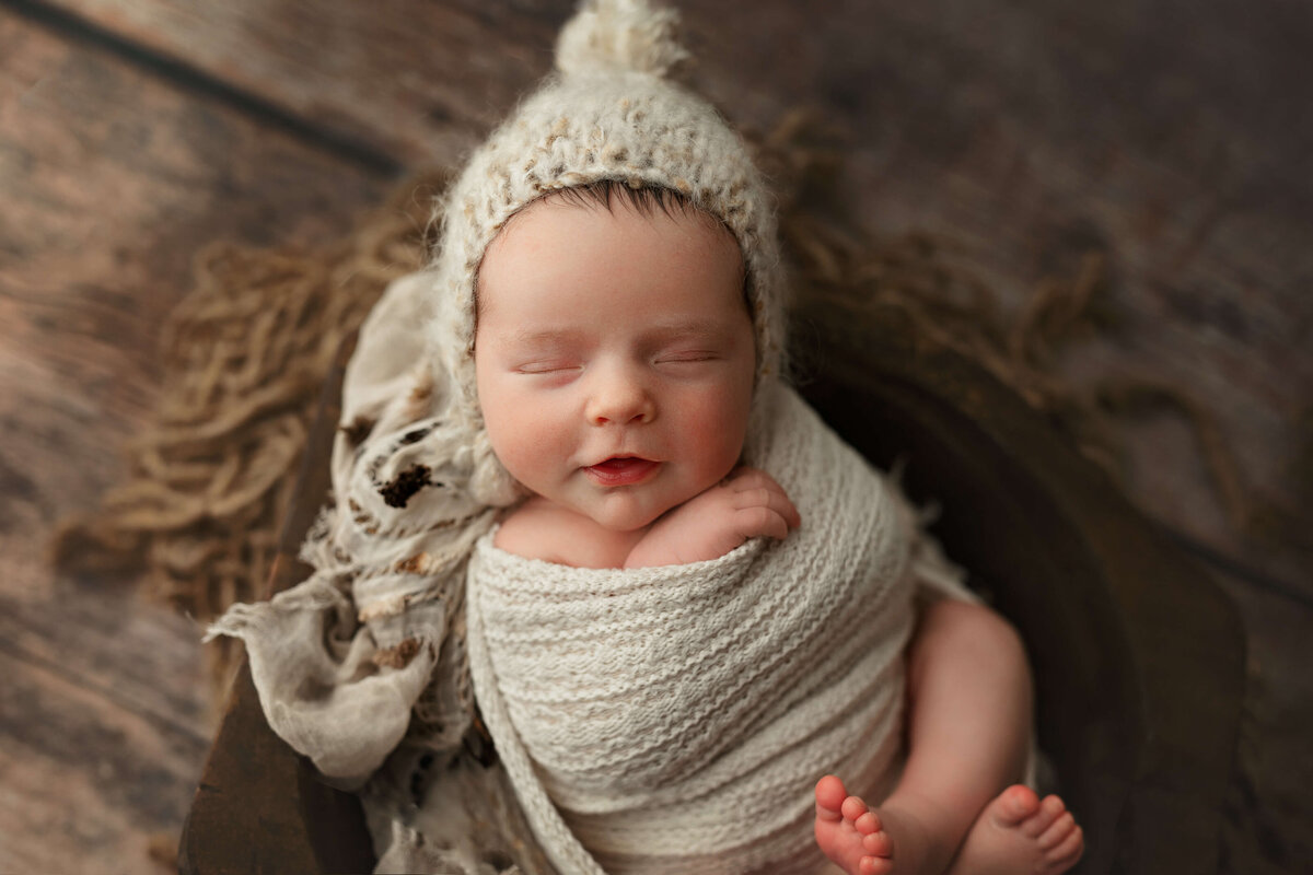 newborn ababy in a pom pom bonnet laying in a wooden bowl at a baby photo shoot
