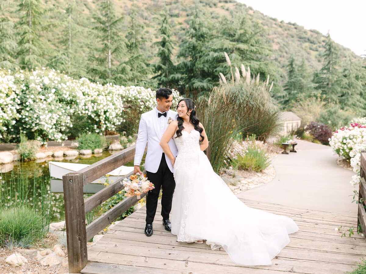 A bride and groom stand on a wooden bridge surrounded by lush greenery and white flowers. The couple smiles warmly, exuding joy and love.
