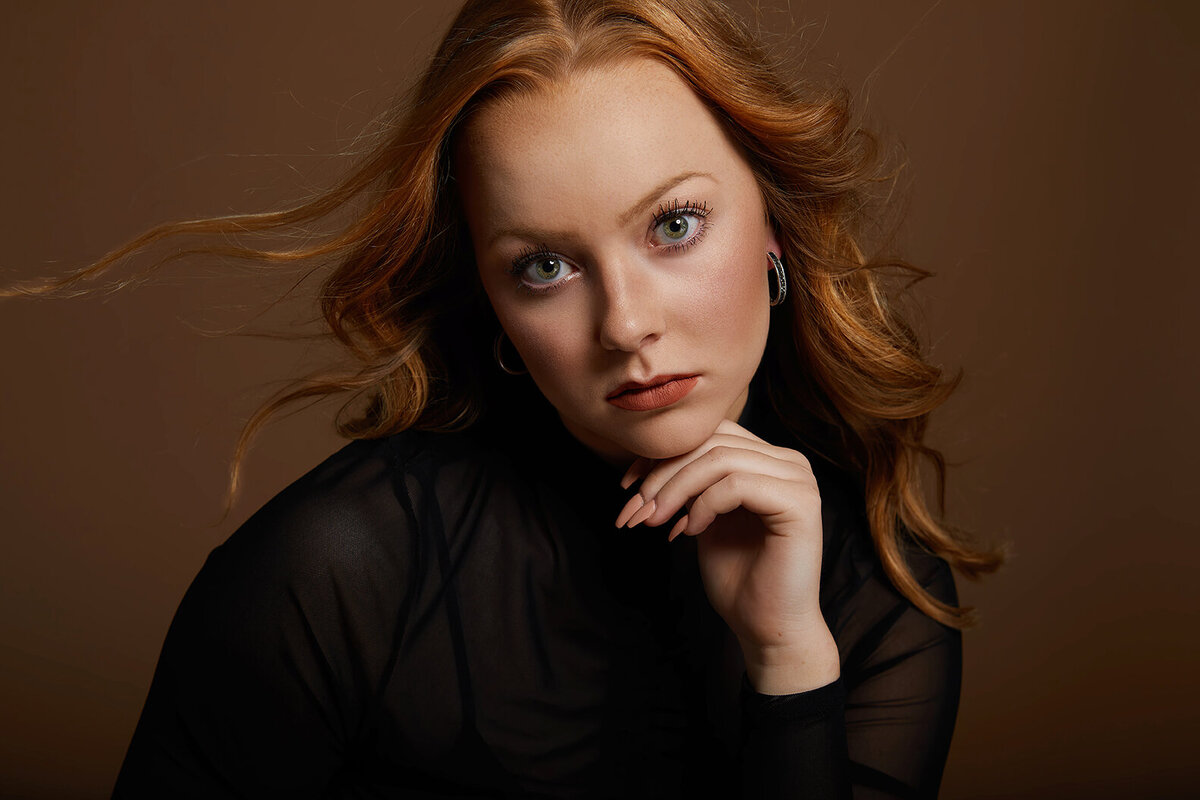 Red hair high school senior girl with hair flying serious pose on brown backdrop.