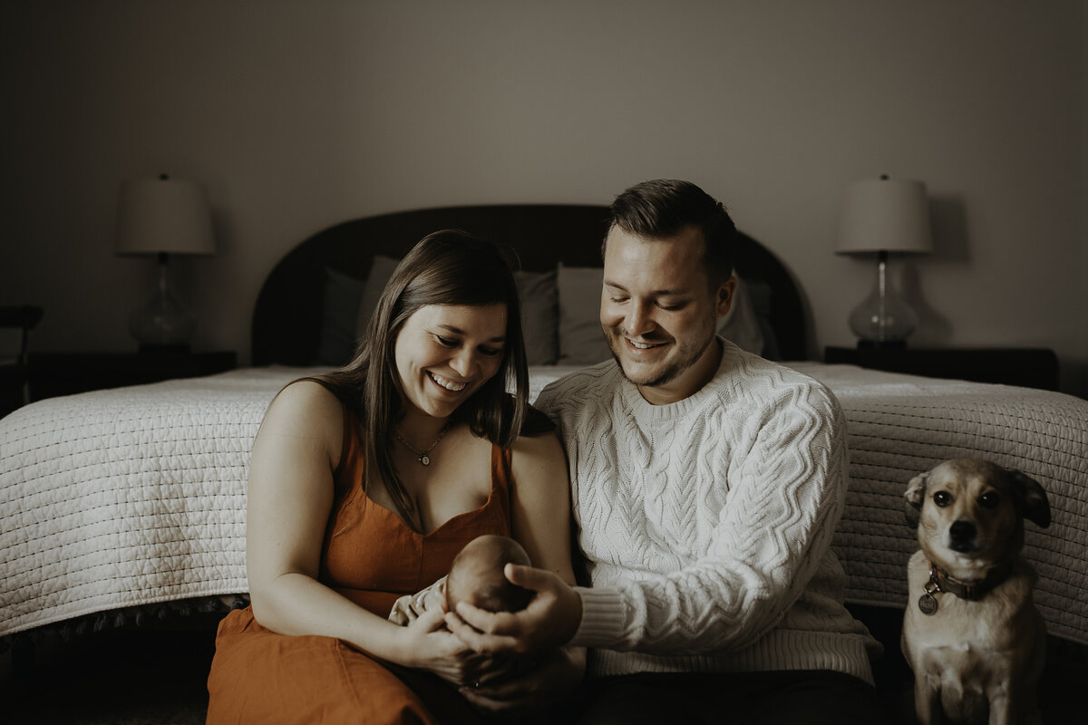 Verona B Photography captures a family sitting on the floor of their bedroom holding their adorable sleeping newborn while the parents gaze tenderly at him and their dog looks at the camera. It is such an honor to capture these intimate family moments at an in-home newborn session near Akron, Ohio.