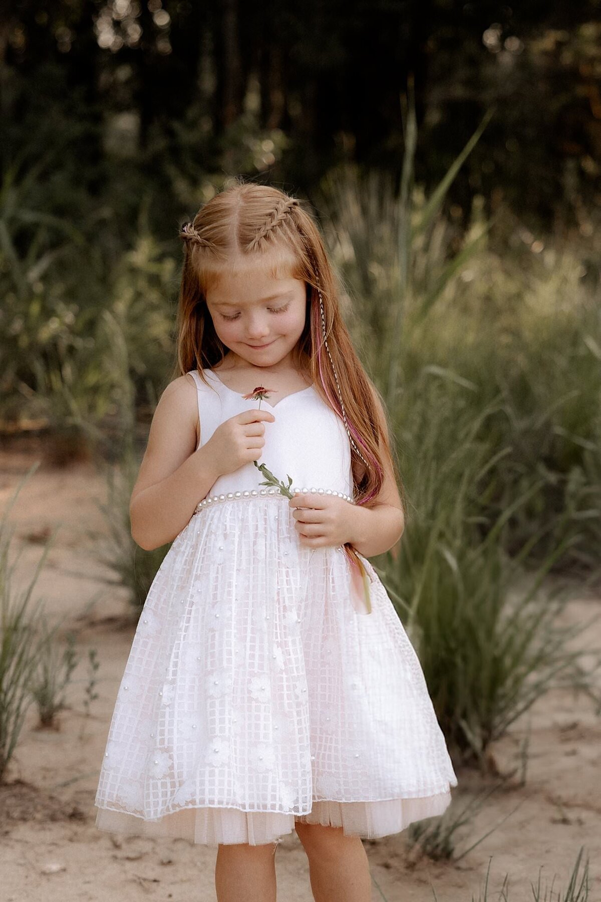 little girl in a white dress holding a flower looking down at the flower in her hand.