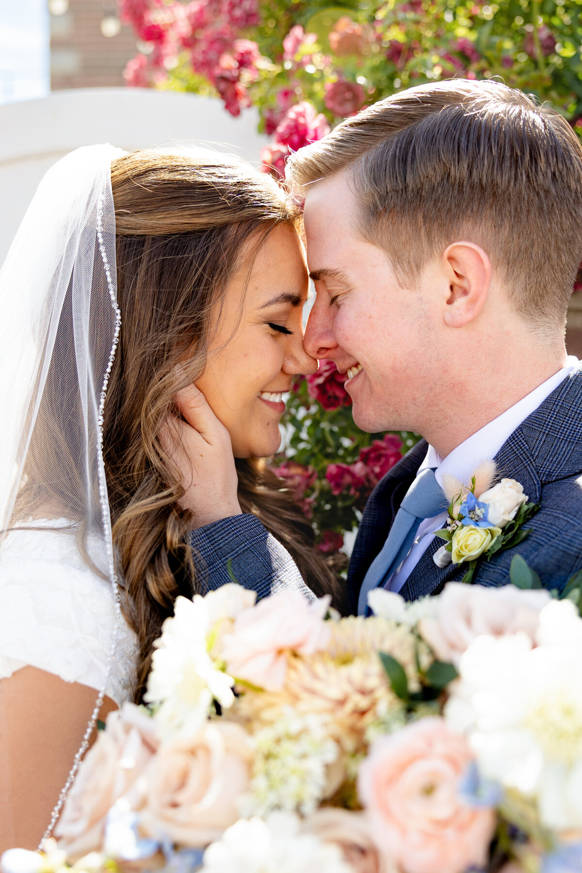 A close-up of a bride and groom touching foreheads and smiling lovingly, surrounded by pink roses.