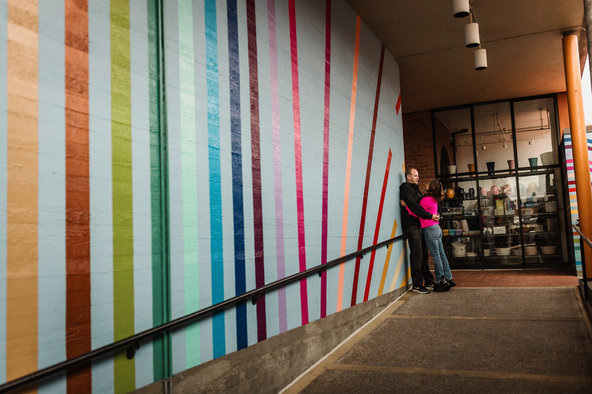 San Francisco couples photography session. Couple stands in front of rainbow stripe wall at Ghiradelli Square