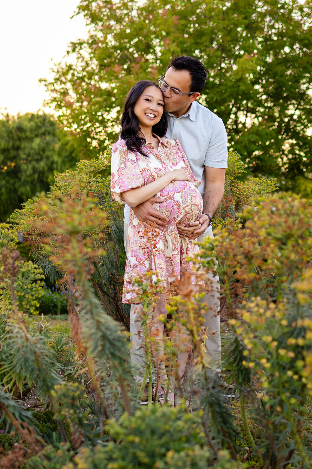 Happy couple stands among native plants as dad-to-be kisses mom’s cheek during their maternity session — Bay Area Maternity Portfolio - Ellobelle Photography.