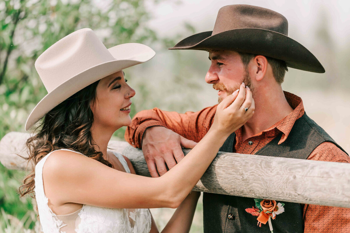 bride and groom lean on a fence and look at each other. Bride touches his face.   