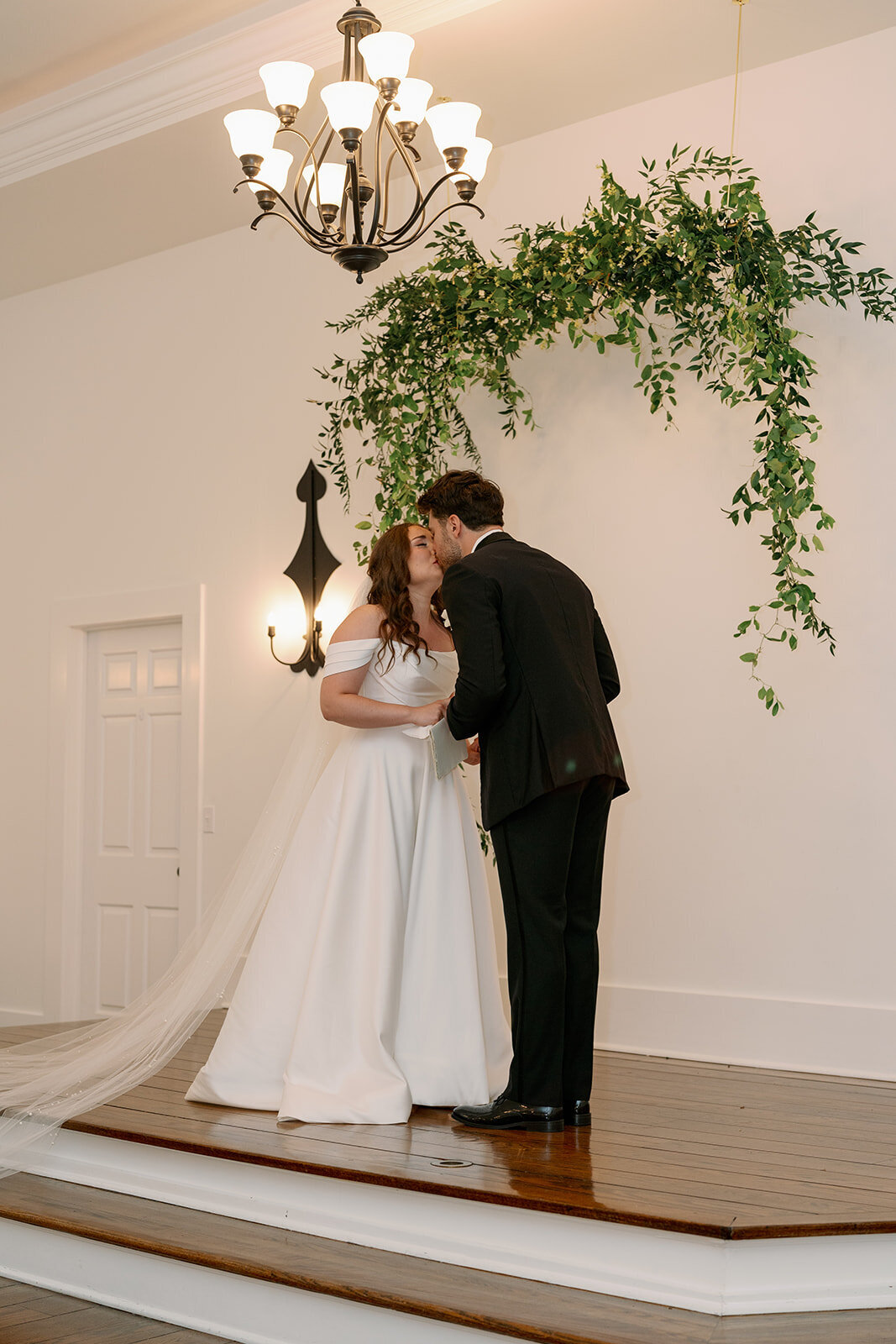 Bride and groom kissing on the ceremony stage at The Morris Estate chapel beneath greenery installation.