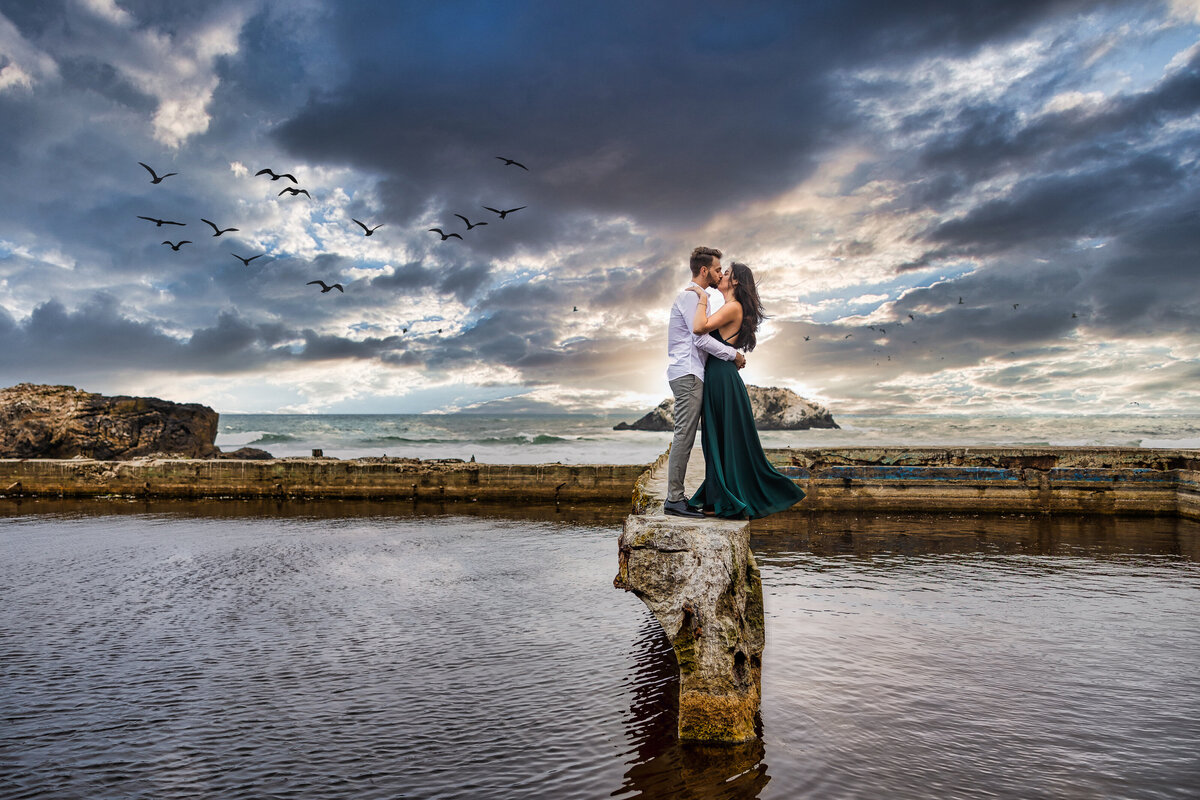 Engaged couple kiss on ledge in front of the San Francisco Bay with clouds in the sky and sunset in the background. Photo by Sacramento wedding photographer, philippe studio pro.
