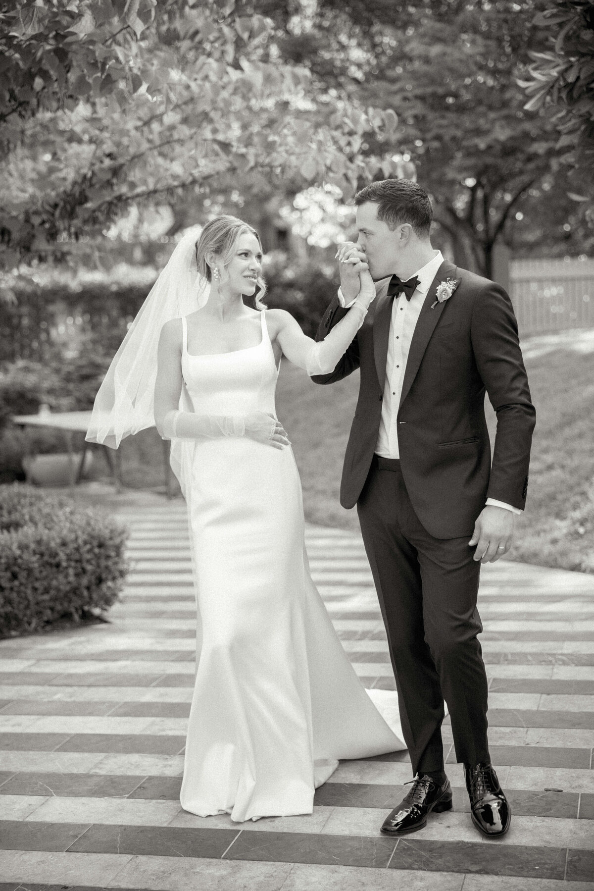 Groom kissing brides hand while walking outside at wedding.