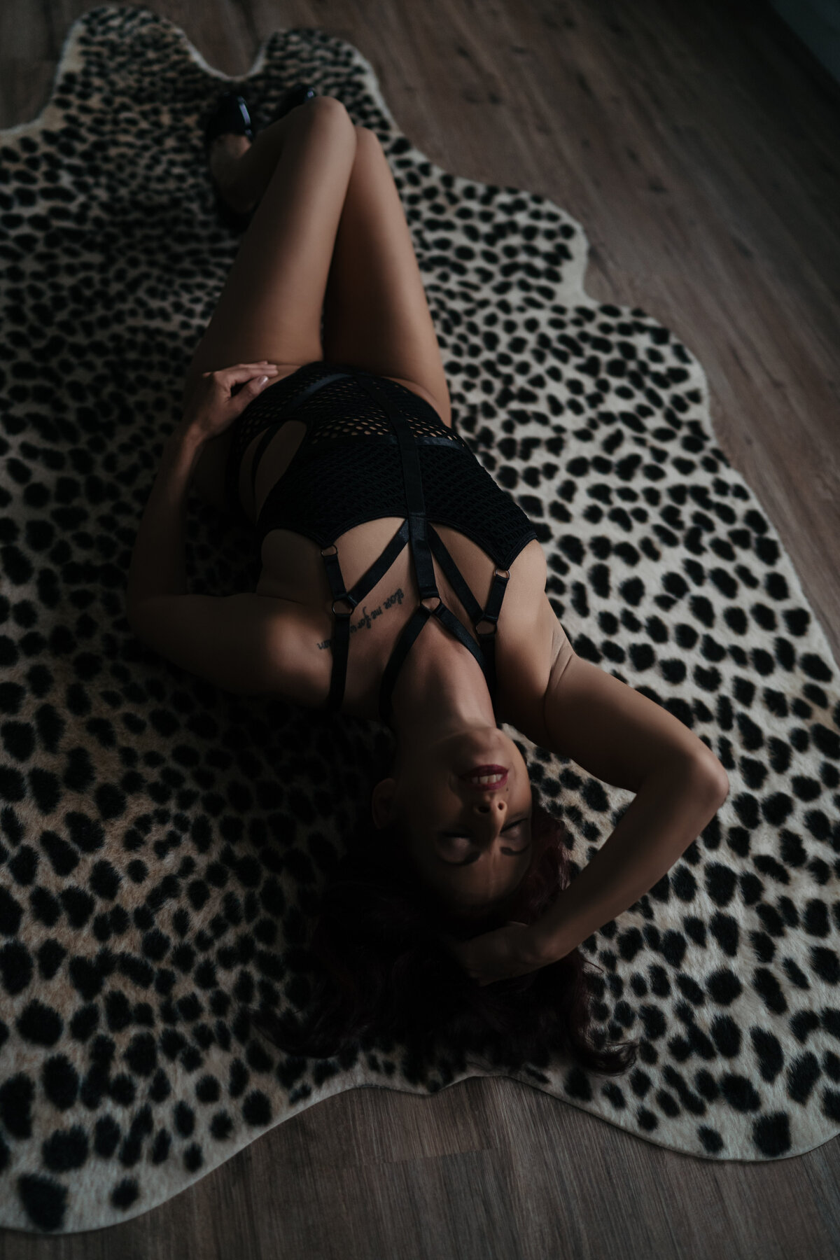 A woman wearing black lingerie arches her back and runs a hand through her hair while laying on a leopard print run on wood floor while posing for a boudoir photography session with Kerry Callahan in Boston