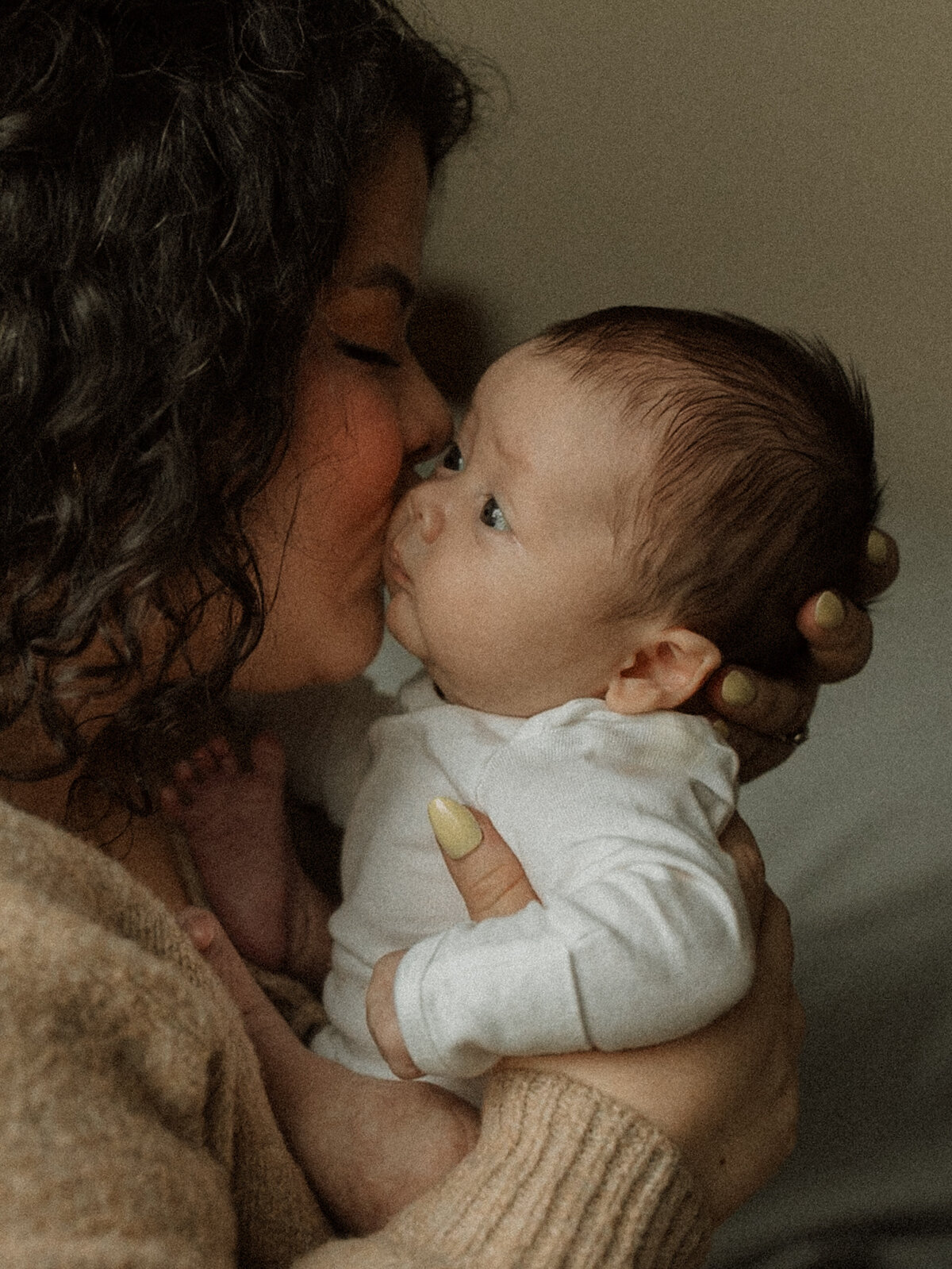 A mother holding her baby close, leaning in for a kiss; the baby gazes out a window in a cozy home setting.