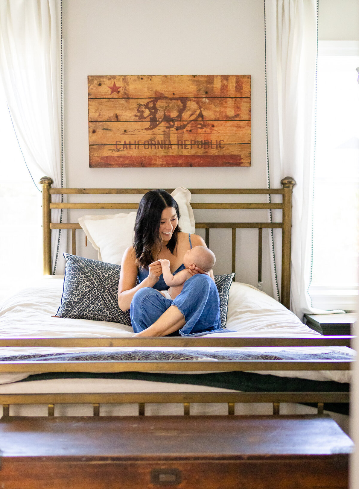 Mother sitting on bed cradling baby beneath California Republic wall art, photographed by San Francisco newborn photographer.