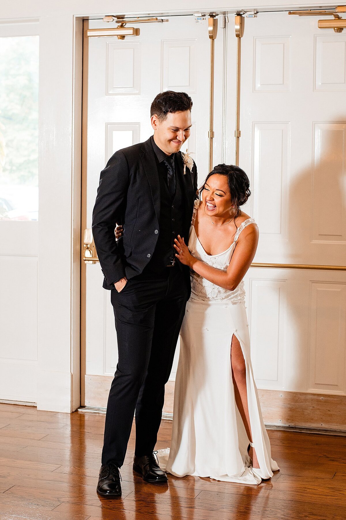 The bride holds onto the grooms arm as they enter their reception laughing together. The bride is wearing a fitted wedding gown with spaghetti straps and a thigh high slit in the skirt. The groom is wearing a black suit with a black shirt and a white rose boutonniere.