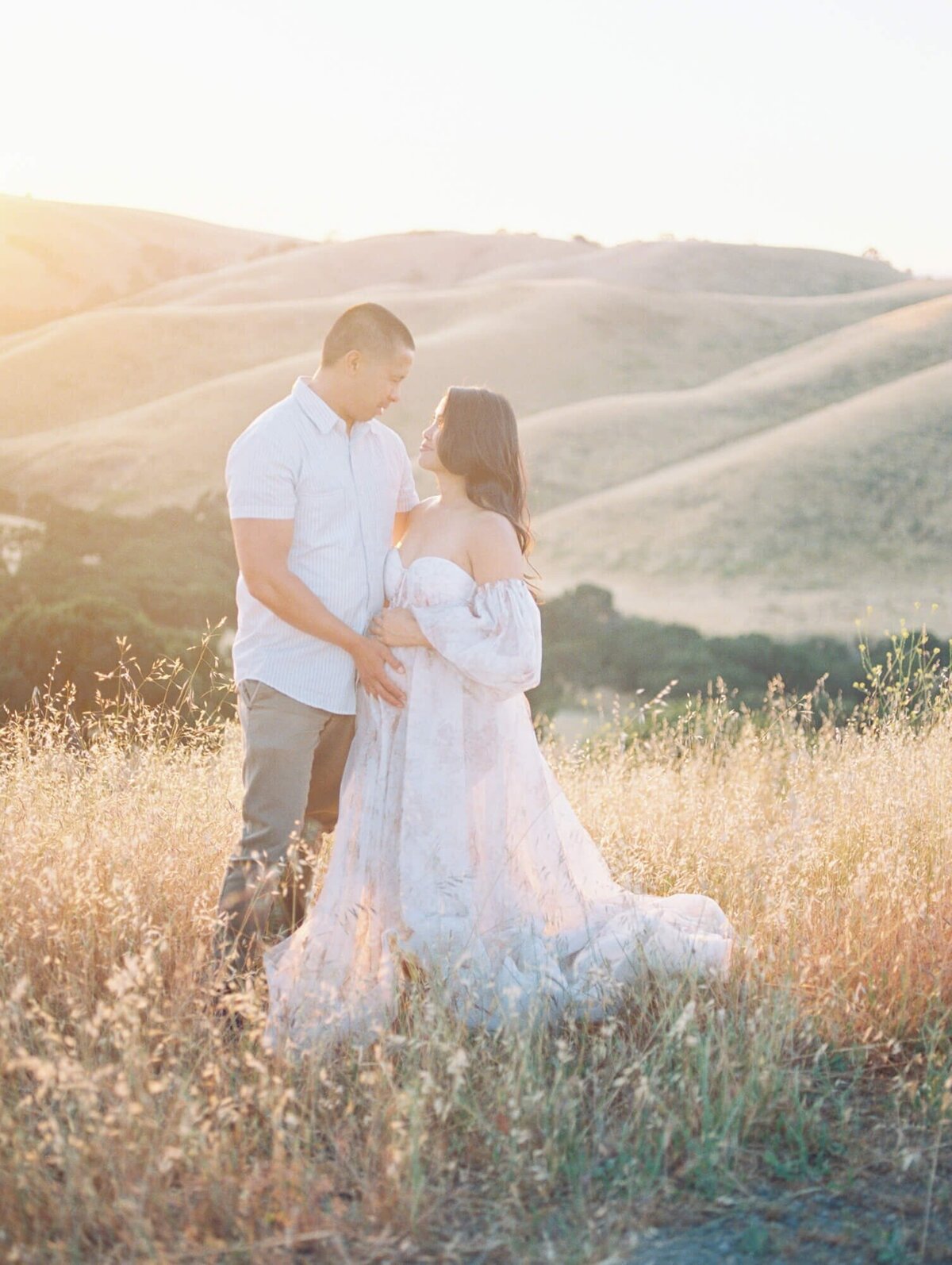 Maternity photoshoot in San Francisco with a pregnant woman and expecting dad standing in a field against hills.