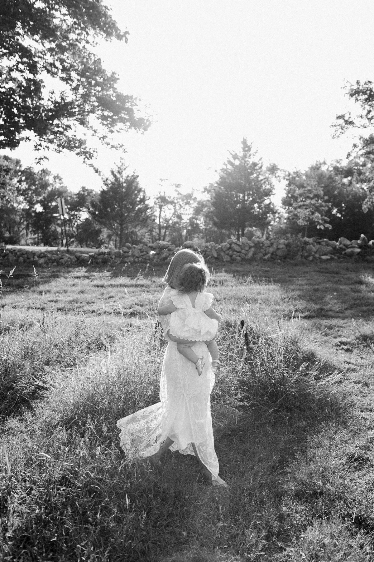 Mother holds child in white dress as she walks through a flower field.